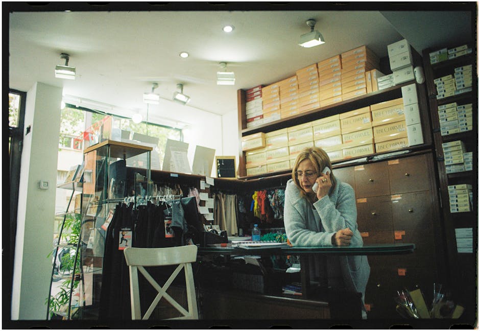 Woman working in a clothing store, talking on the phone near merchandise racks.